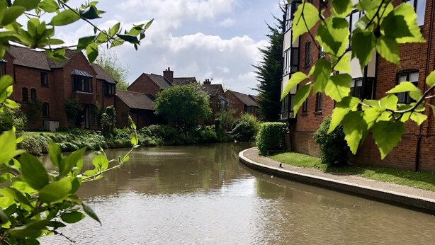 A canal with brick buildings on either side, surrounded by greenery.