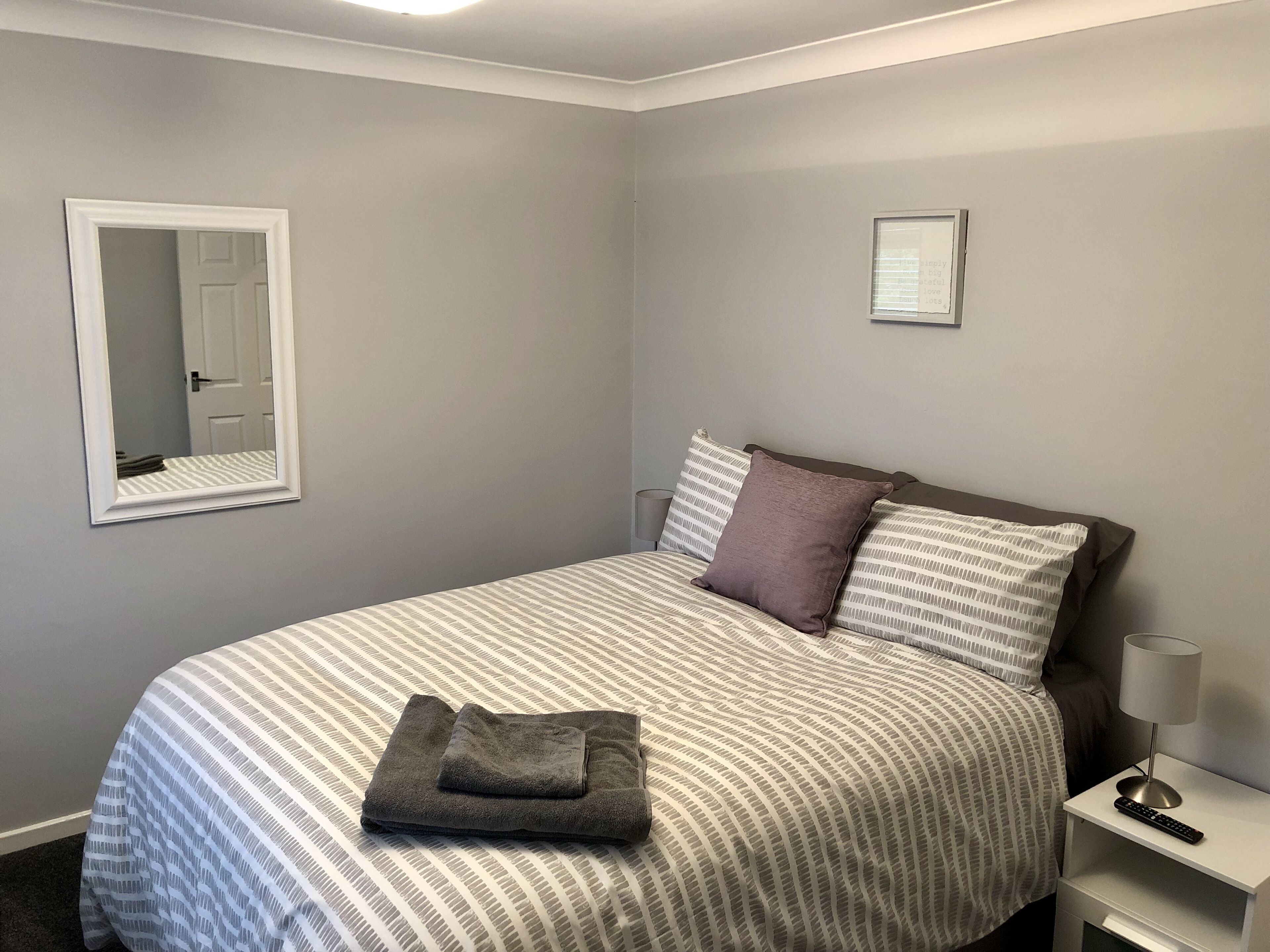 Un lit bien fait, recouvert d’une couverture à rayures, avec une serviette pliée et deux oreillers. On y trouve une table de chevet avec une lampe et une télécommande.
