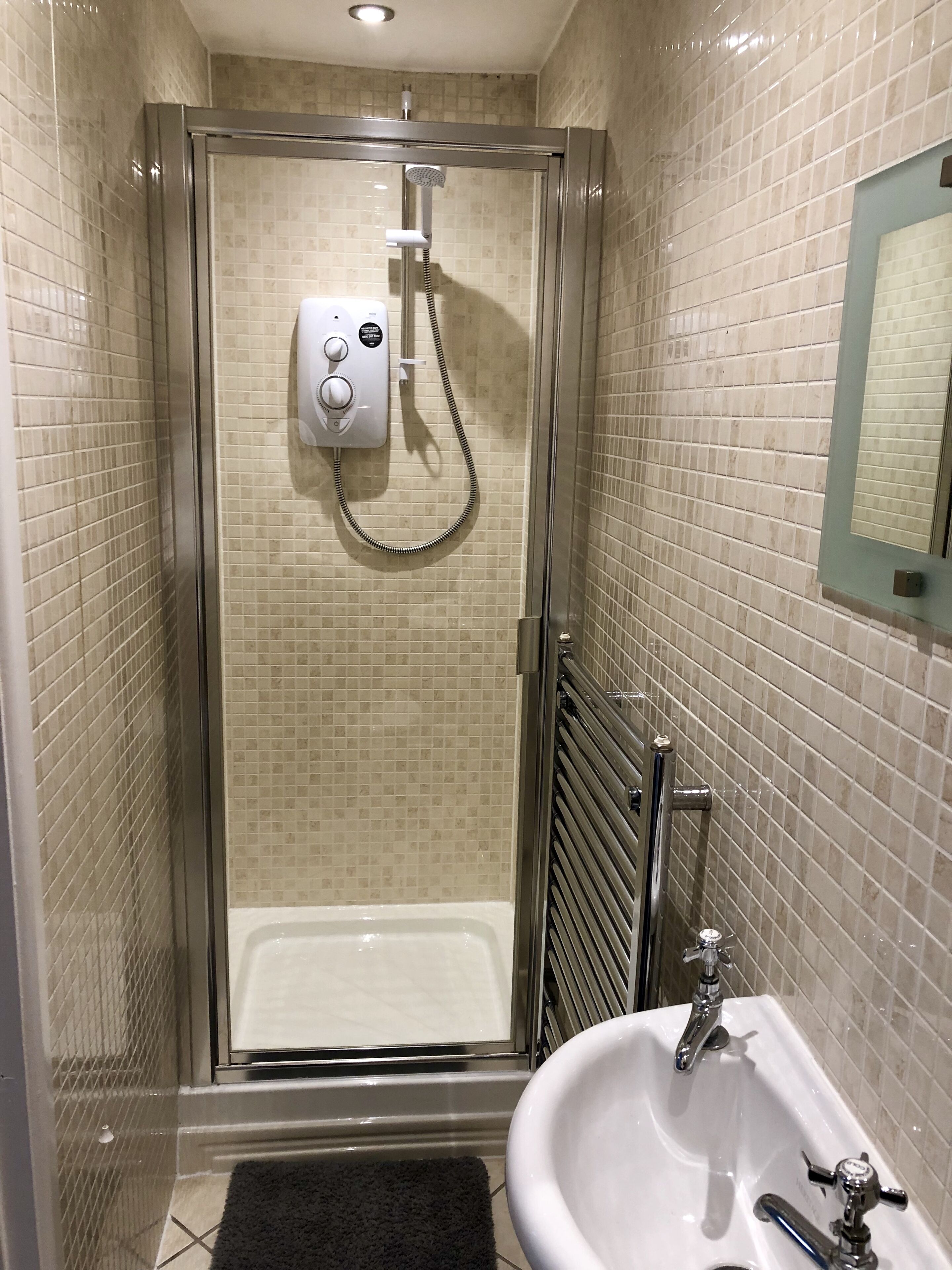 A glass-enclosed shower with a handheld showerhead and control panel, a white sink with a single faucet, and a tiled wall with a mirror.