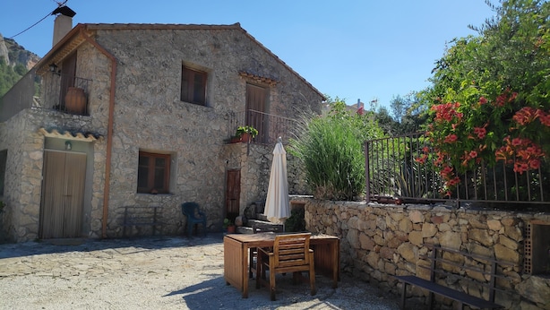A stone building with a balcony, a wooden table and chairs, and a stone wall with a bench.