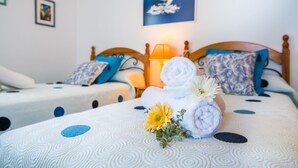 Ein Hotelzimmer mit zwei Betten, jedes mit einer gemusterten Tagesdecke, Kissen und einem zusammengerollten Handtuch. Auf dem Bett befinden sich eine Blume und eine Lampe.