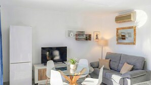 Ein modernes Wohnzimmer mit grauem Sofa, weißen Sesseln, einem Glastisch, einem Flachbildfernseher, einem weißen Kühlschrank und einer Wand-Klimaanlage.