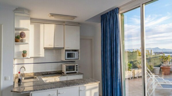 A modern kitchen with white cabinets, a granite countertop, a microwave, and a sink. There is a window with blue curtains, and a view of a balcony with plants and a distant landscape.