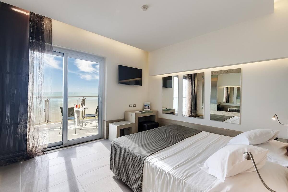 A modern hotel room with a large bed, a sitting area with a table and chairs, a balcony with a view, and a flat-screen TV mounted on the wall.