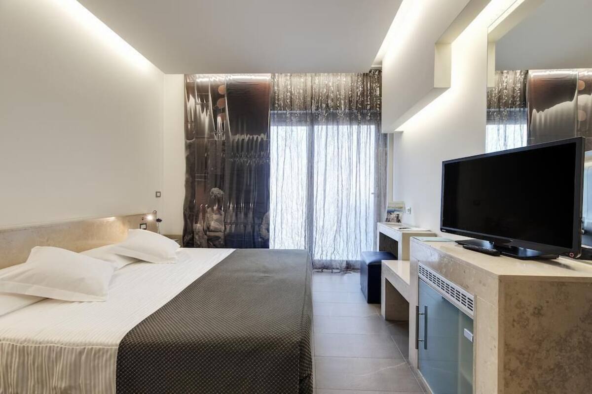 A modern hotel room with a large bed, a flat-screen TV, a desk with a chair, and a large window with curtains.