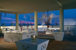 A modern outdoor lounge area with white sofas, a glass table, and decorative plants.