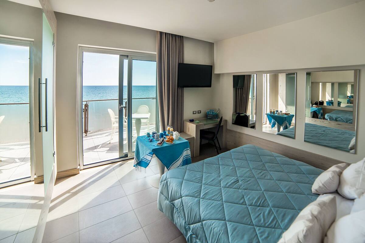 A hotel room with a bed, a desk, a chair, a table with breakfast items, a TV, and a balcony with a view of the sea.