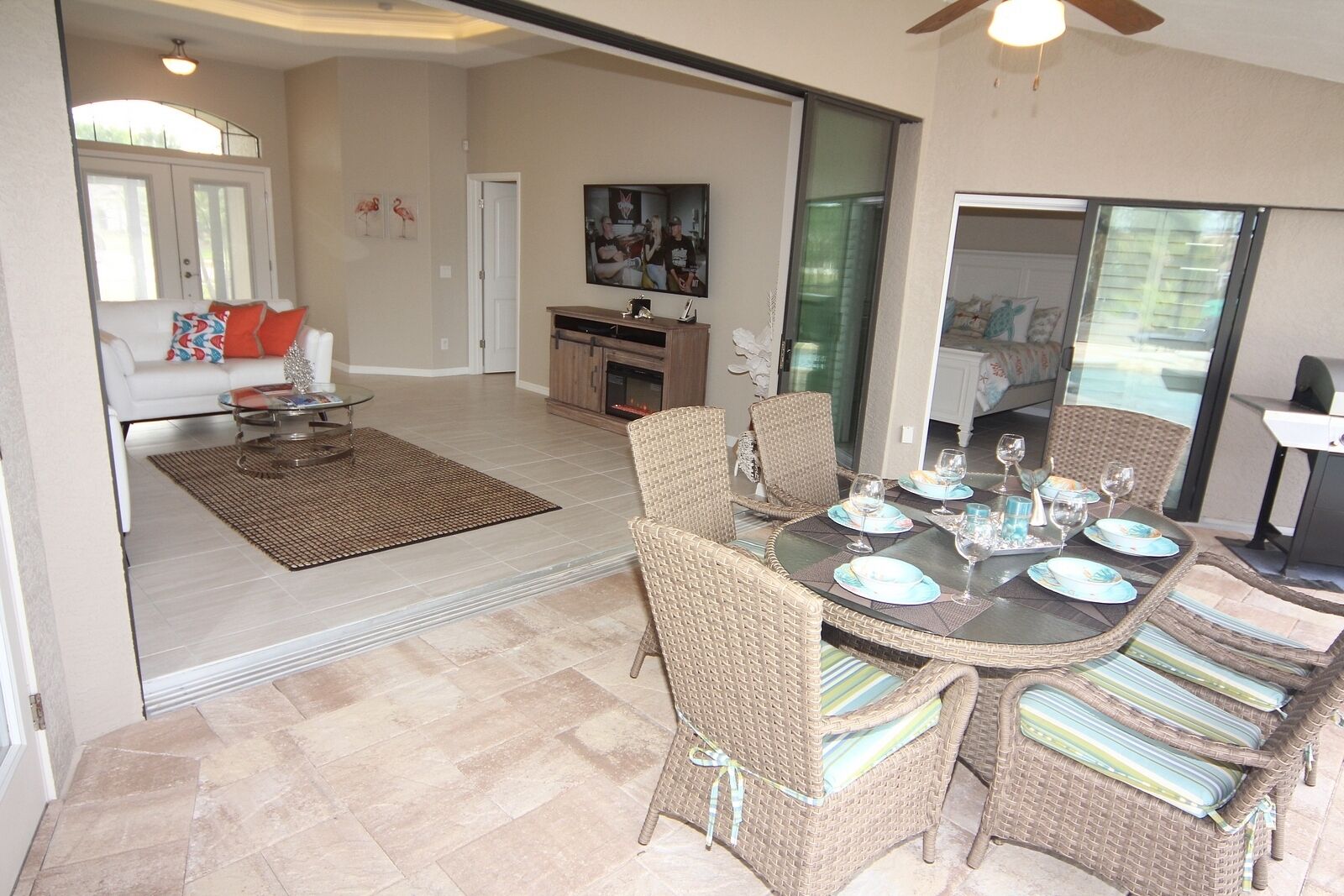 A spacious living area with a dining table set for four, surrounded by wicker chairs with striped cushions. A kitchen with a large island and a dining area with a glass door leading outside are visible.