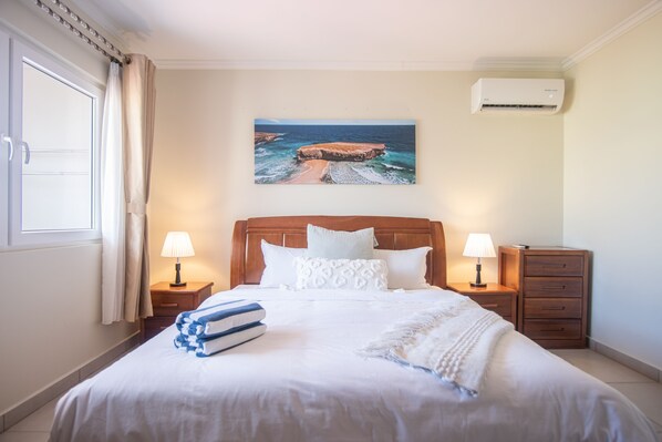 A neatly made bed with white linens and a blue and white towel stack, two bedside lamps, a wooden nightstand, a small drawer, and a painting of a beach scene on the wall. - 10% discount for weekly stay. 2 Bedrooms 2 Baths Condo in Eagle Beach Aruba! (Noord)