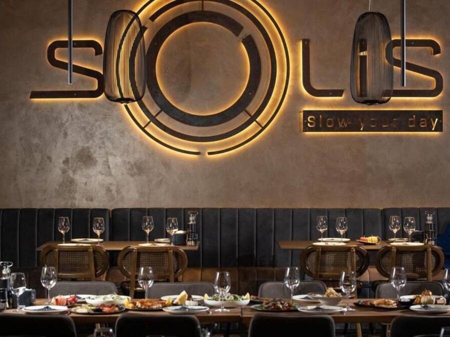 L’intérieur d’un restaurant au design moderne, avec une enseigne murale portant le nom « Solis » et le slogan « Prenez le temps de savourer ». Les tables sont dressées avec des verres à vin, des assiettes et des couverts, et des banquettes confortables sont disposées le long du mur.
