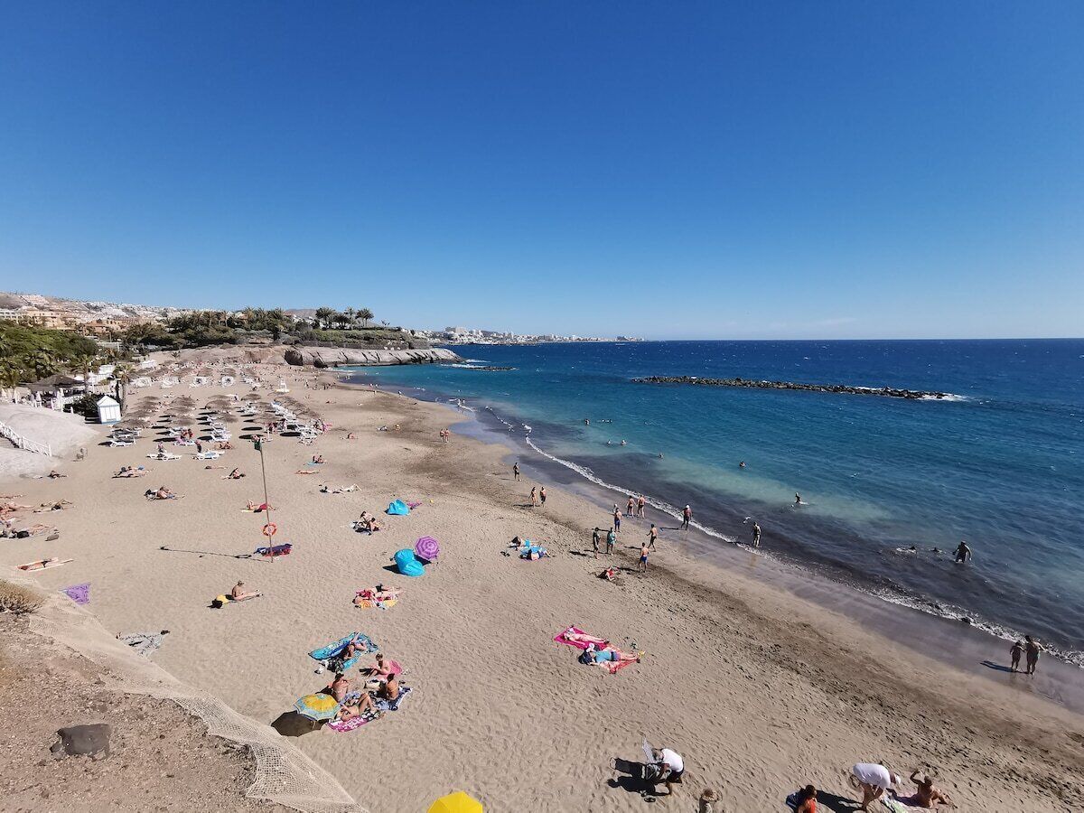 Ein Strand mit badenden und sonnenden Menschen, blauem Himmel und einer felsigen Küste.