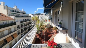 A balcony with a view of neighboring buildings, potted plants, and a table with flowers. - Lovely balcony in Musiciens, central Nice. (Nice)