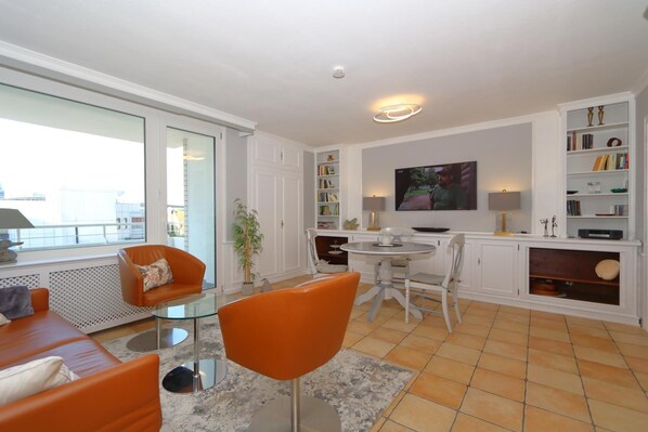 A modern living room with a glass coffee table, orange chairs, a white fireplace, a TV mounted on the wall, and a bookshelf.