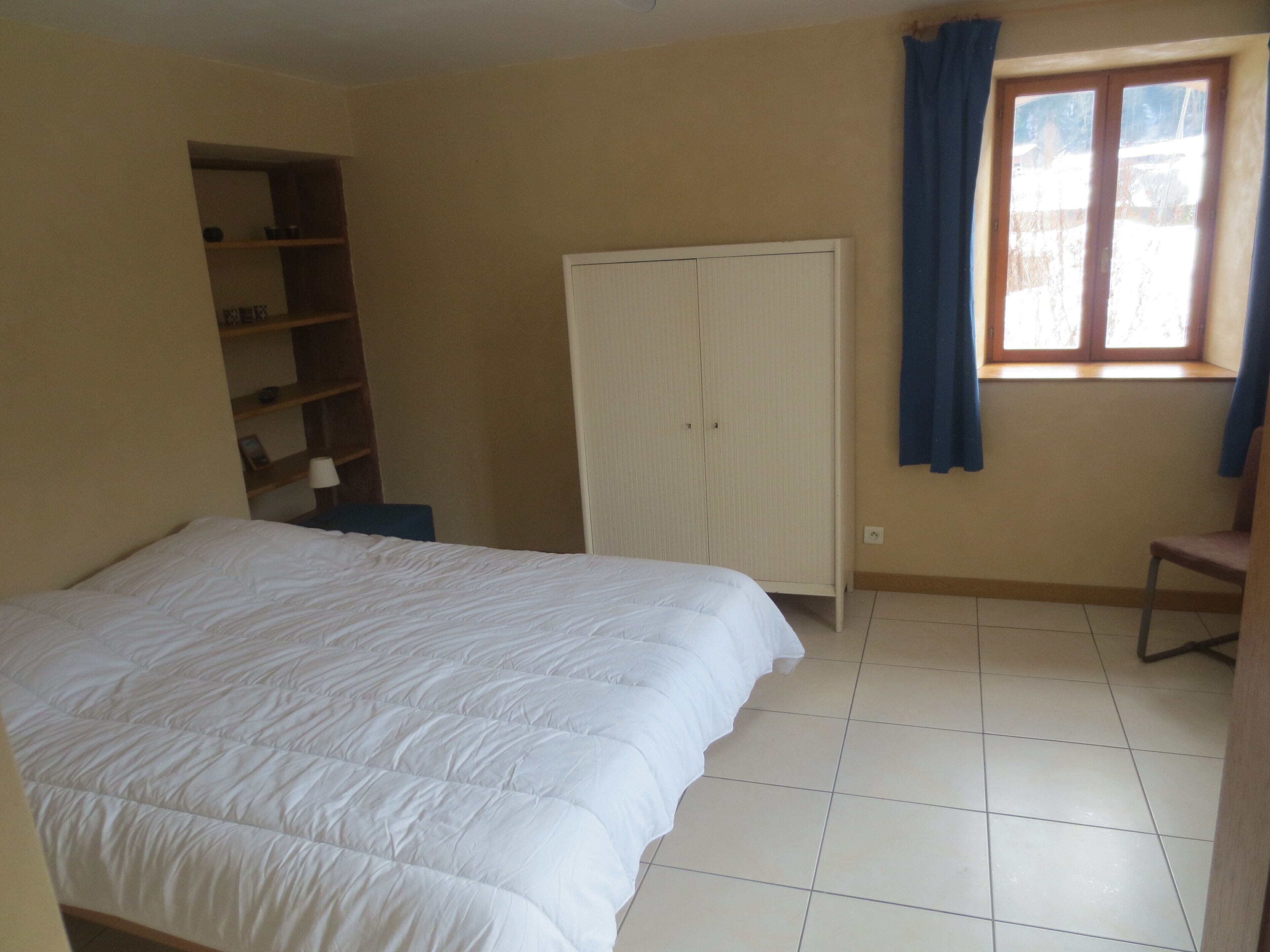 A bedroom with a bed, a wardrobe, a window with curtains, and a shelf unit.
