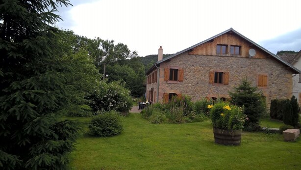 Un bâtiment en pierre, doté d’un toit en pente et de volets en bois, entouré d’un jardin bien entretenu où poussent diverses plantes et fleurs.