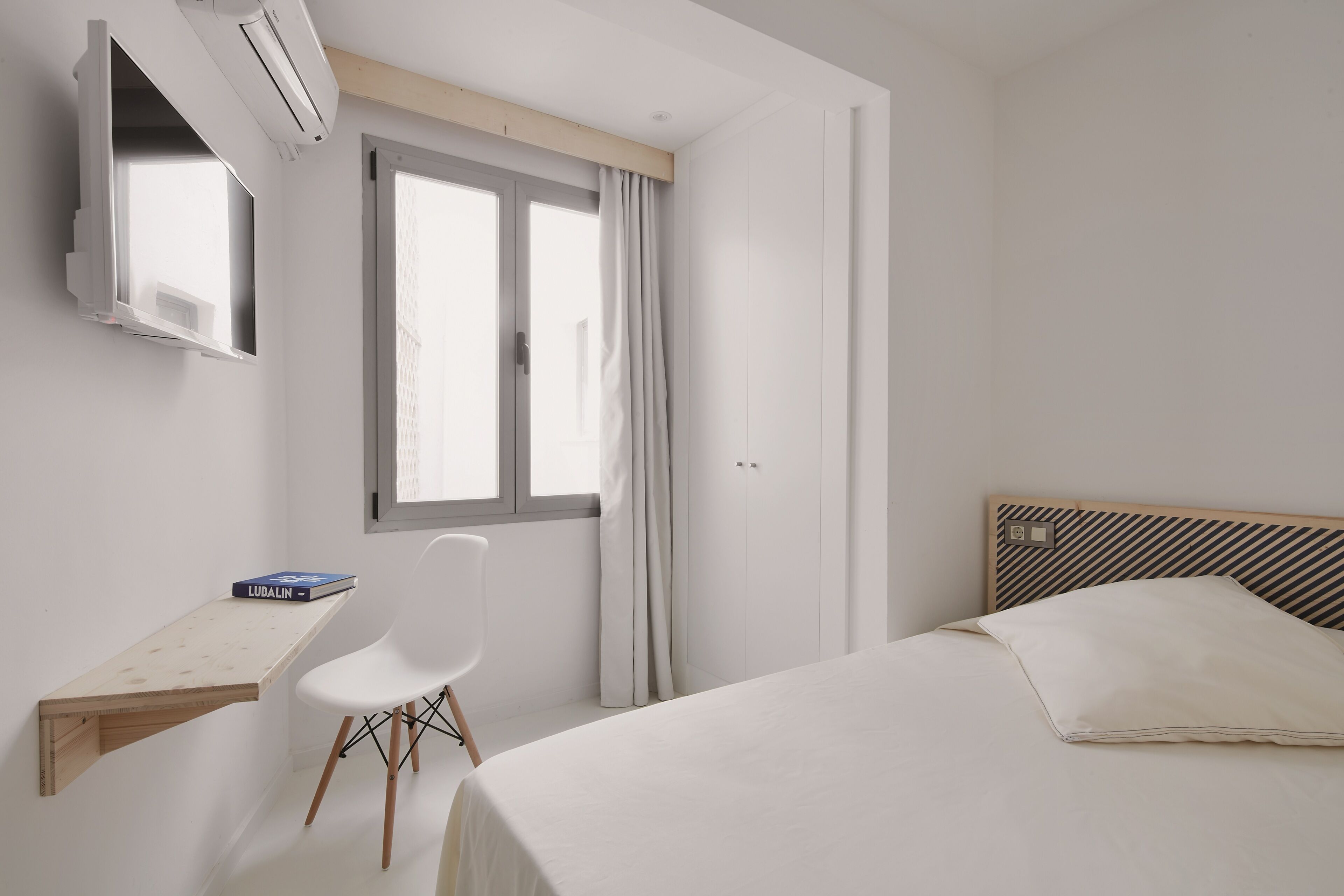 A bedroom with a bed, a desk, a chair, a TV, and a window.