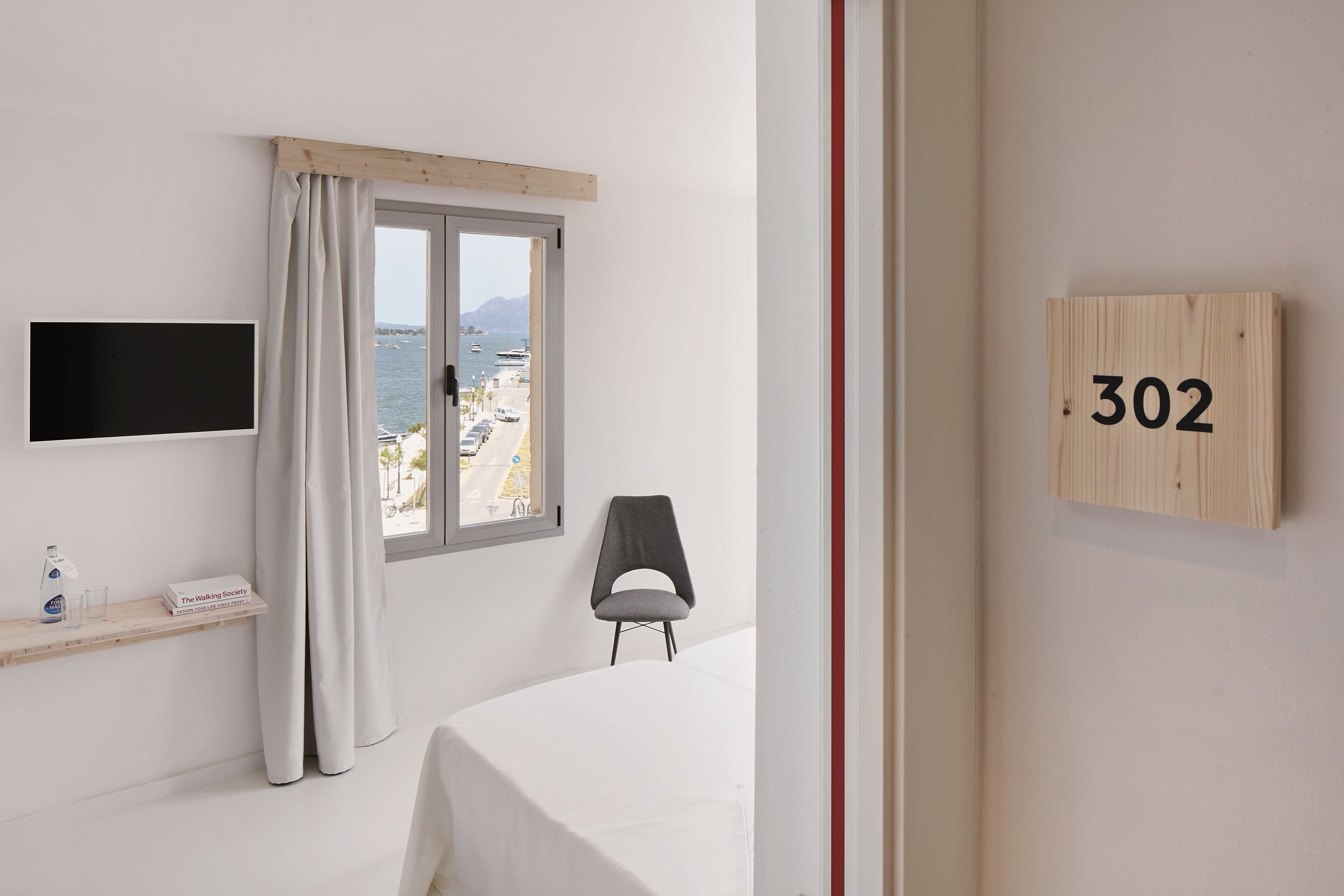 A hotel room with a bed, a TV mounted on the wall, a window with curtains, a chair, and a wooden sign with the number 302.