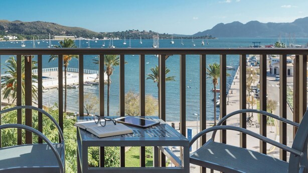 A balcony with a table, chairs, and a view of the sea with boats and a distant town. - Eolo Affiliated by FERGUS (Pollenca)