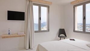 A hotel room with a bed, a chair, a TV mounted on the wall, a window with a view of the sea, and a shelf with a bottle and glasses. - Eolo Affiliated by FERGUS (Pollenca)