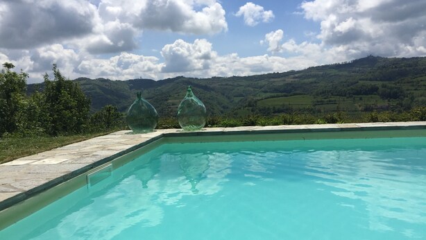 A swimming pool with clear blue water, surrounded by a stone deck. In the background, there are green hills and a cloudy sky. - Authentic stone house in the heart of Piedmont - with pool and wild views (Cessole)