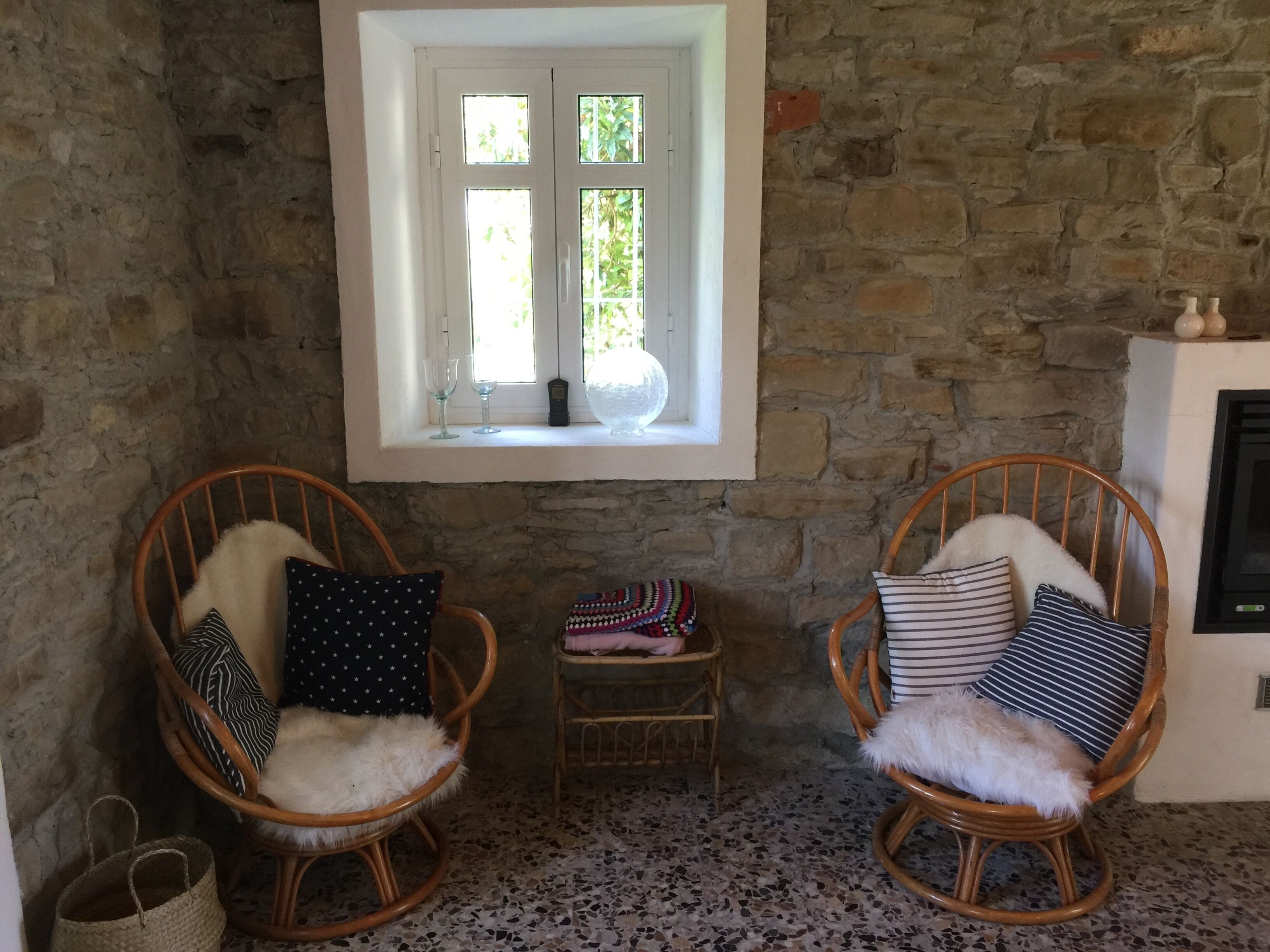 Une pièce chaleureuse aux murs de pierre, deux fauteuils à bascule en osier dotés de coussins moelleux, une petite table surmontée d’un panier tressé et une fenêtre donnant sur un paysage verdoyant.