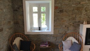 A cozy room with stone walls, two wicker rocking chairs with fluffy cushions, a small table with a woven basket, and a window with a view of greenery. - Authentic stone house in the heart of Piedmont - with pool and wild views (Cessole)