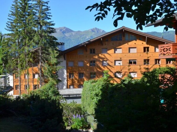 A multi-story building with a wooden facade, surrounded by greenery and mountains in the background.