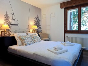 Ett hotellrum med en säng, två sänglampor, en stol, ett fönster med utsikt över träd och en väggmålning som föreställer en skidlift.