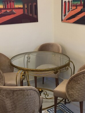 Un comedor con una mesa de vidrio rodeada de sillas con cojines, un tapete estampado y dos cuadros en la pared.