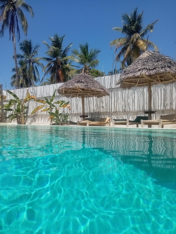 Una piscina cristalina con palmeras y sombrillas de paja al fondo.