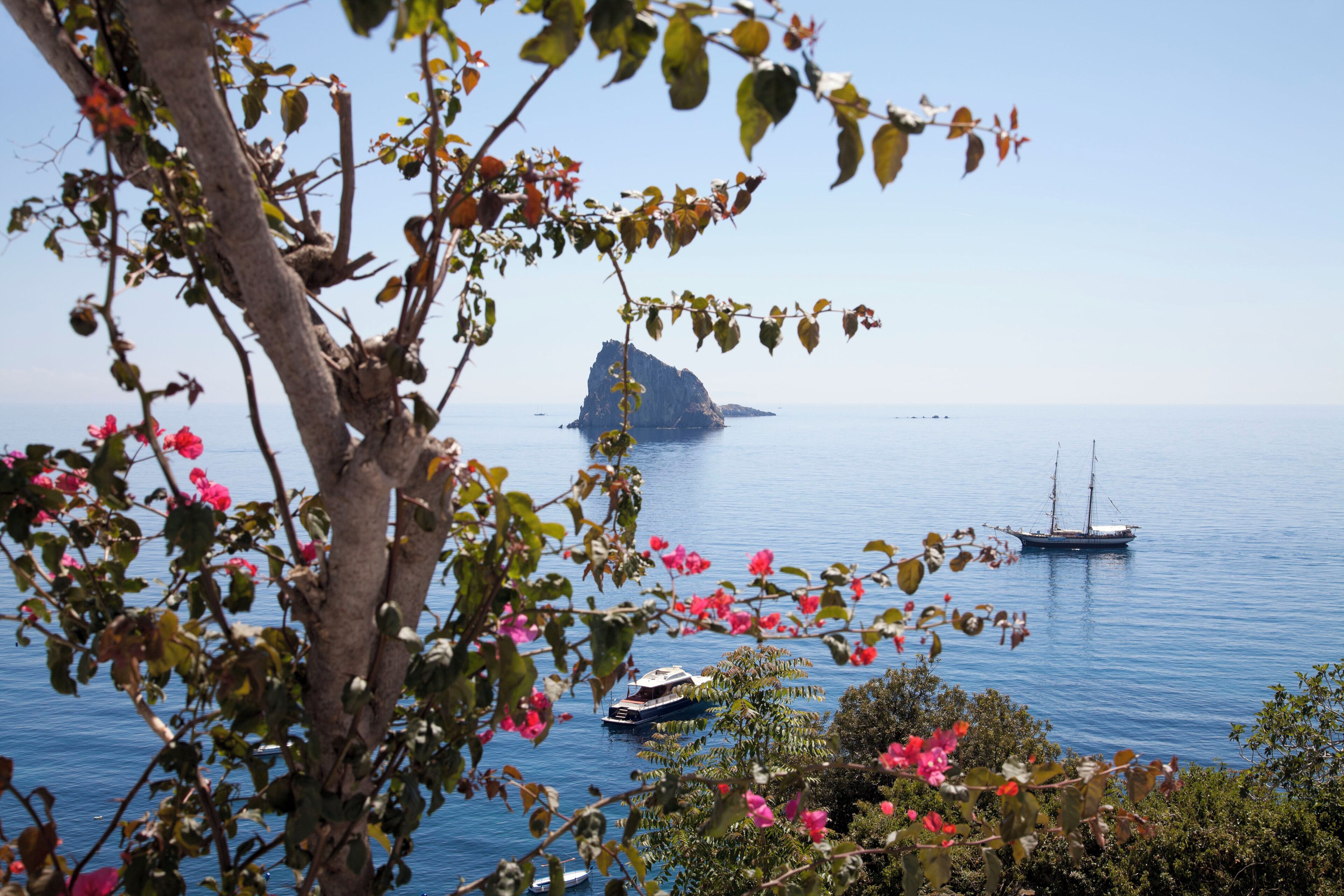 A coastal scene with a large rock formation in the distance, boats on the water, and lush greenery in the foreground.