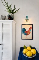 Una pared con un cuadro colorido de un gallo enmarcado, una planta de aloe en maceta y un cuenco de limones sobre una mesa azul.