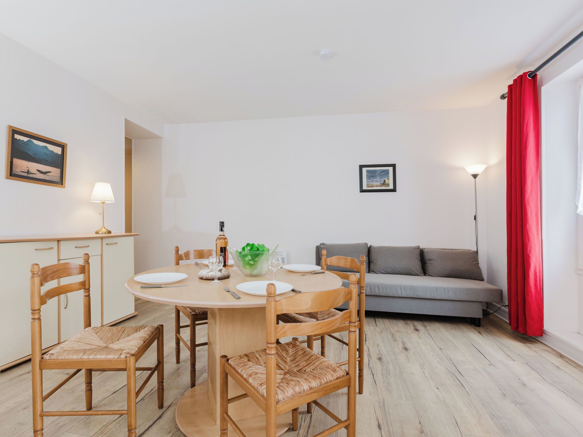 A dining area with a round table, chairs, a bottle, and a bowl of greens.