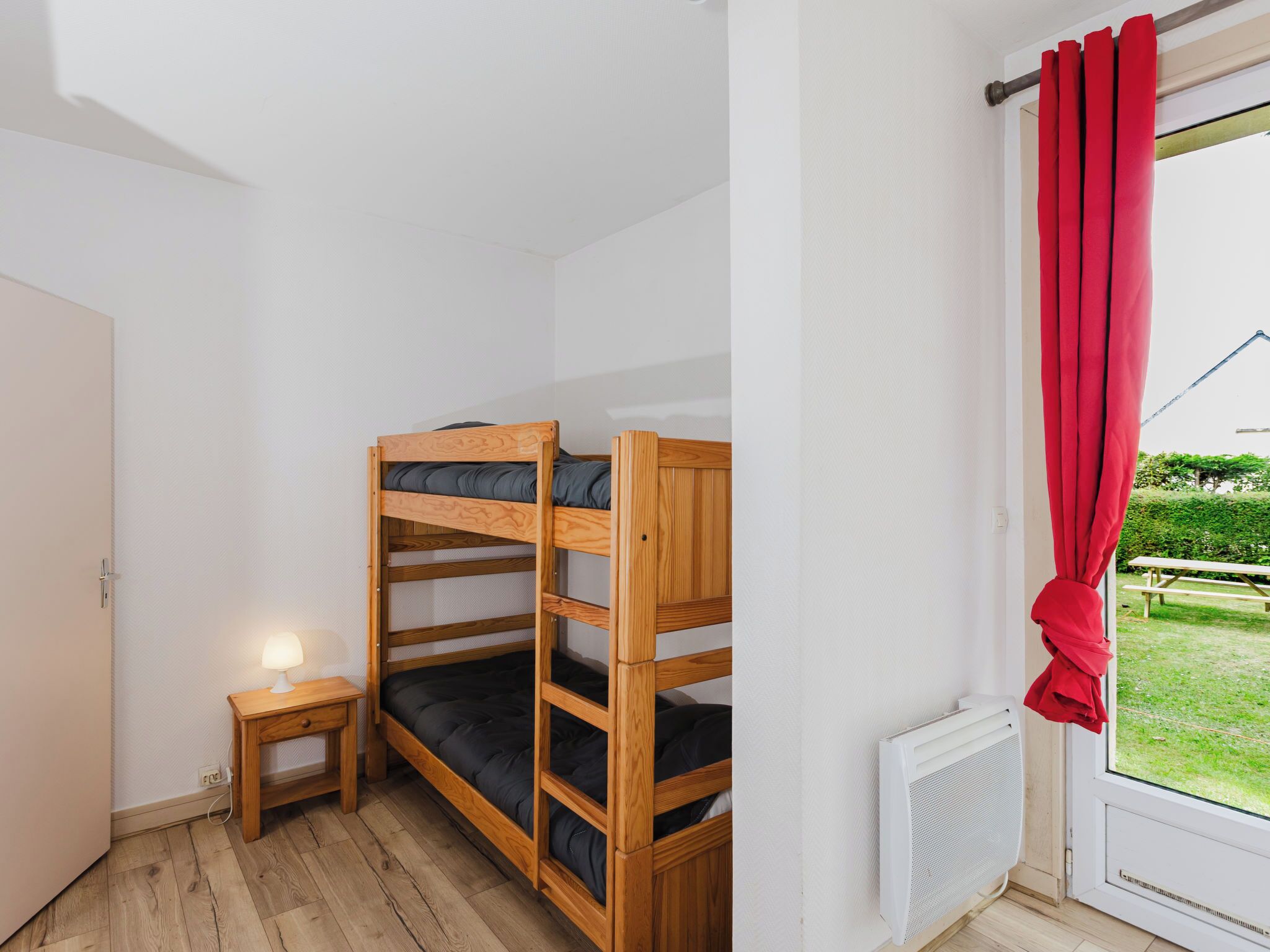 Ein Zimmer mit einem Etagenbett, einem kleinen Nachttisch mit Lampe, einem Fenster mit roten Vorhängen und einem Heizkörper.