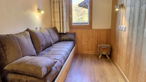 A cozy room with a brown sofa, a small stool, a window with a view of a mountainous landscape, and wooden walls.