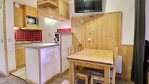A compact kitchen with wooden cabinets, a built-in microwave, a red-tiled backsplash, a dining table with chairs, and a flat-screen TV mounted on the wall.