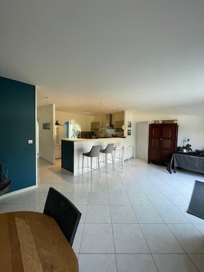 A spacious living area with a kitchen in the background, a dining table with chairs, and a wooden cabinet.