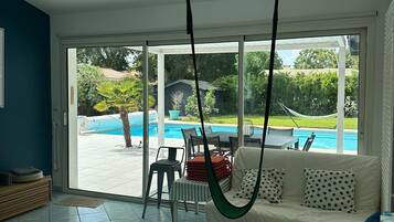 A room with a large sliding glass door leading to a pool, a hanging chair, and a sofa with patterned cushions.