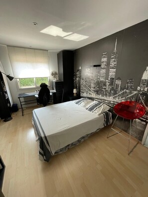 A modern bedroom with a large bed, a desk, and a cityscape mural on the wall.