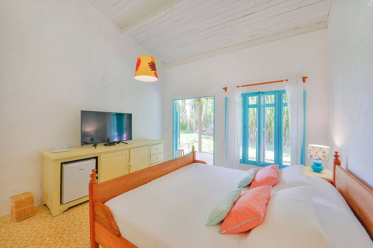 Un dormitorio con cama, televisión, refrigerador pequeño y ventana con persianas azules.