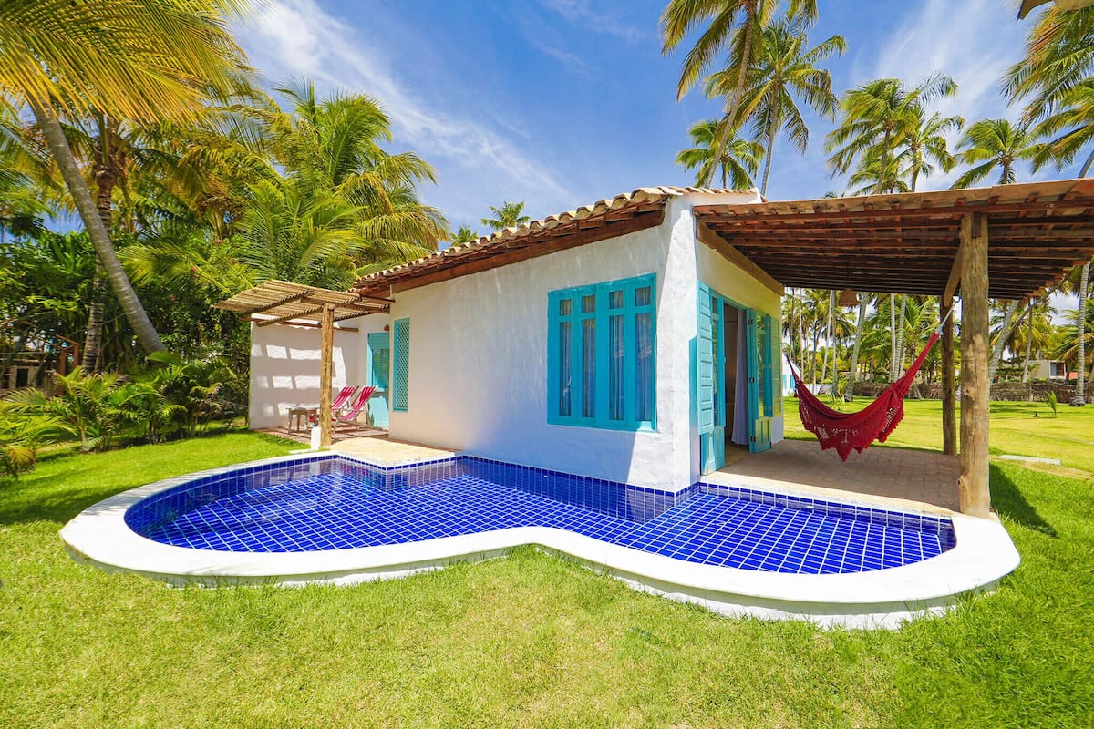 Una piscina con revestimiento de azulejos azules, una pequeña estructura anexa con una hamaca roja, y un césped verde exuberante con palmeras al fondo.