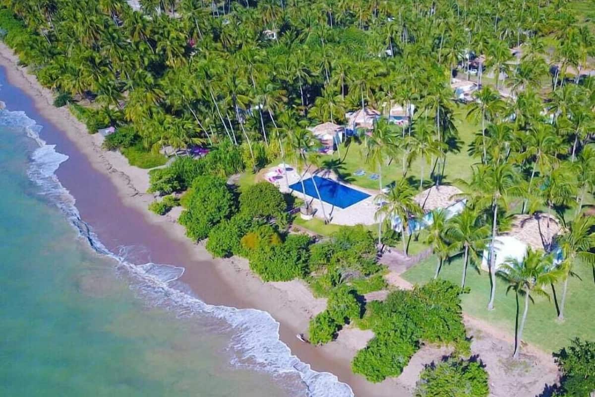 Vista aérea de un complejo turístico en la playa, con piscina, palmeras y propiedades frente al mar.
