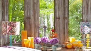 A tropical setting with a wooden window frame, a table with orange juice, candles, and purple flowers. - Pousada Patacho (Porto de Pedras)