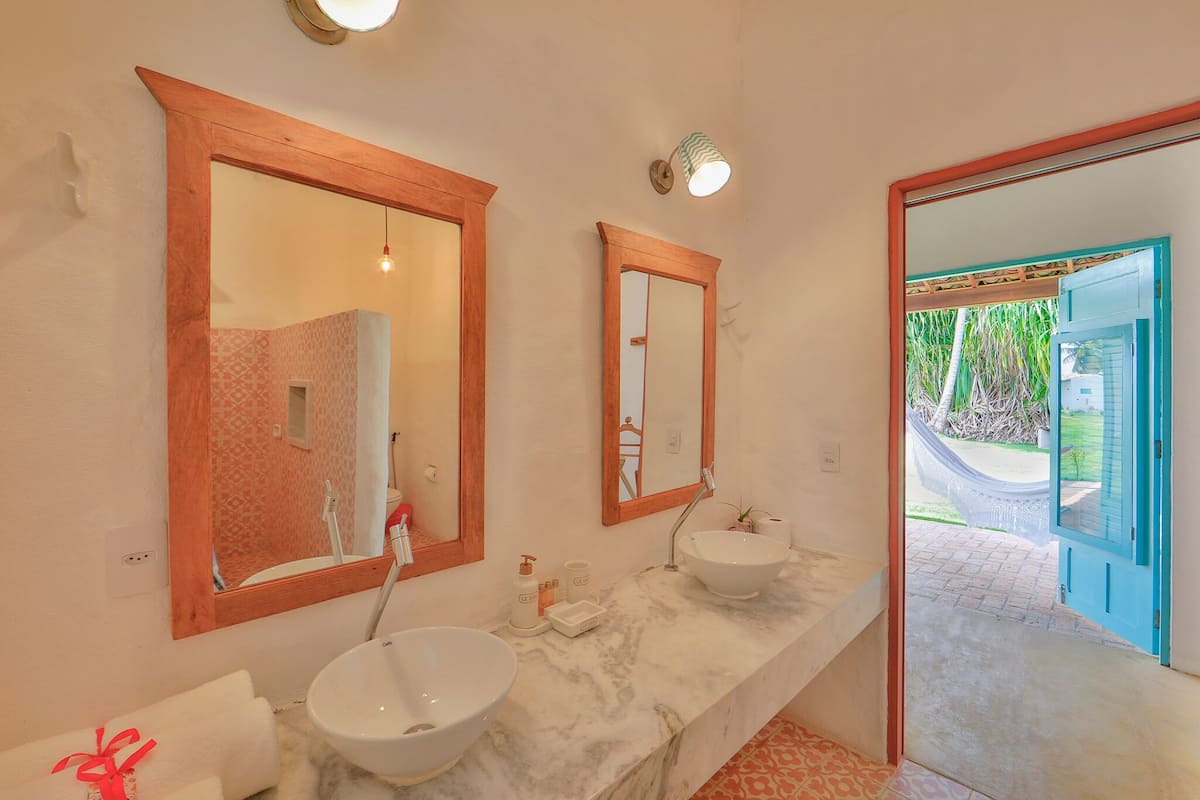 Un baño con dos lavamanos, un espejo grande y vista a un jardín por una puerta abierta.