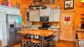 A rustic kitchen with wooden walls, a central island with stools, and a refrigerator. - Deep Creek Waterfront Home • Hot Tub • Pool Table • Minutes to Downtown & GSMNP (Bryson City)
