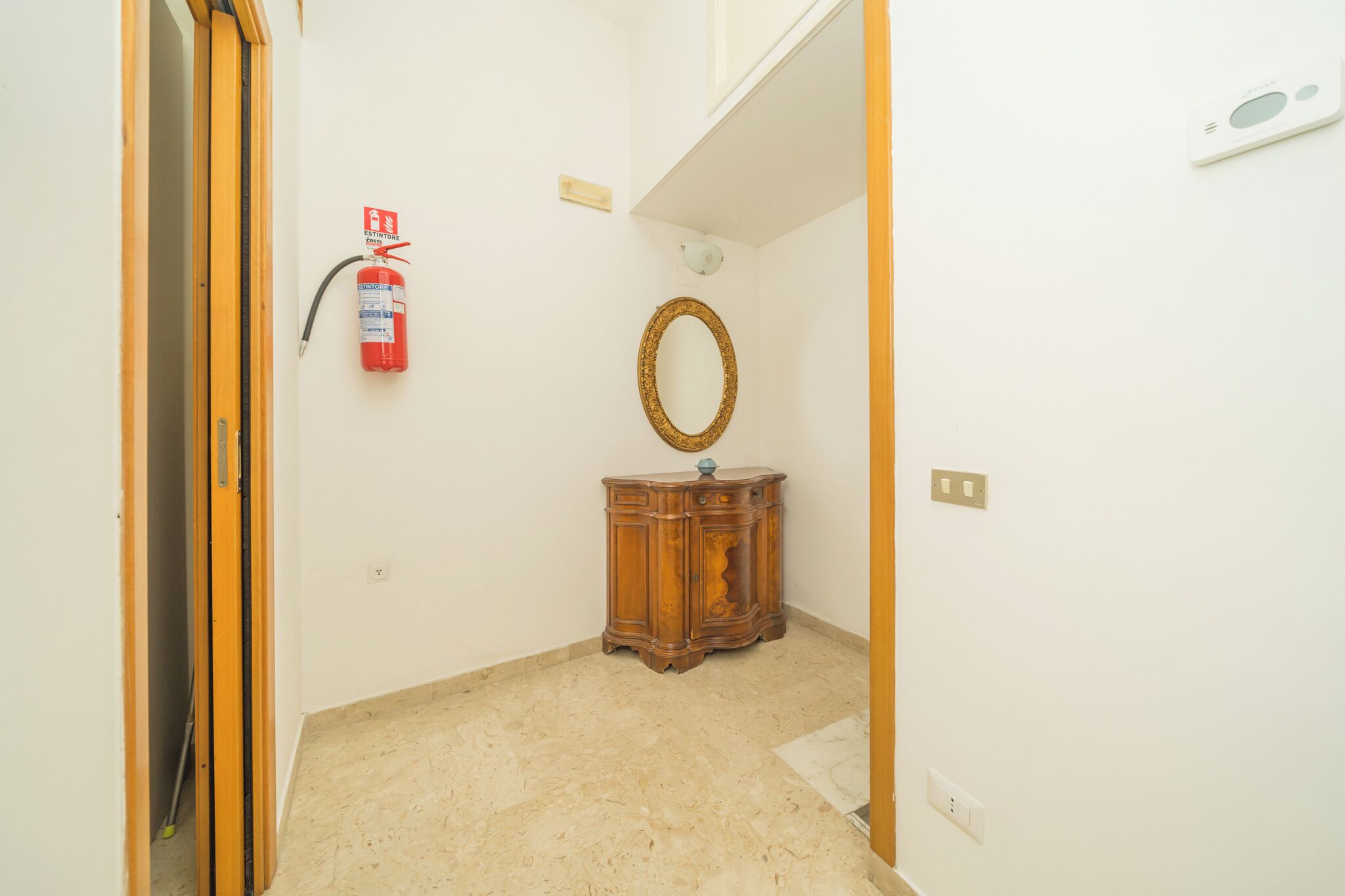 A room with a wooden cabinet, a wall-mounted mirror, a fire extinguisher, and a thermostat.