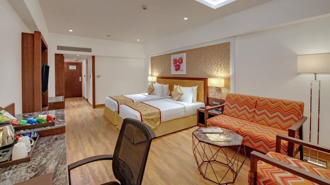 A hotel room with a bed, a sofa, a small table, and a chair. There is a television and a tray with items on the floor.