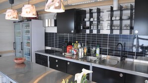A modern kitchen with black cabinets, a stainless steel countertop, and a built-in oven. There are hanging pendant lights, a refrigerator, and a sink with a faucet. - Ecochique (Heuvelland)