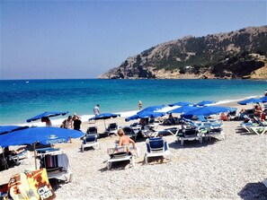 A beach with rows of sun loungers and umbrellas, a rocky hillside in the background, and a clear blue sea. - Vast and beautiful villa  on the Costa Blanca, with magnificent panoramic views. (Altea la Vella)