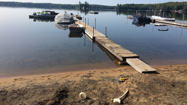 A calm lake with a wooden dock extending into the water, surrounded by boats and a forested shoreline. - Sandy Beach Cottage on Chandos Lake (Apsley)